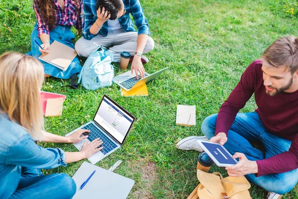 Partial view of multiethnic students with notebooks and digital devices sitting on green grass in park — Stock Photo