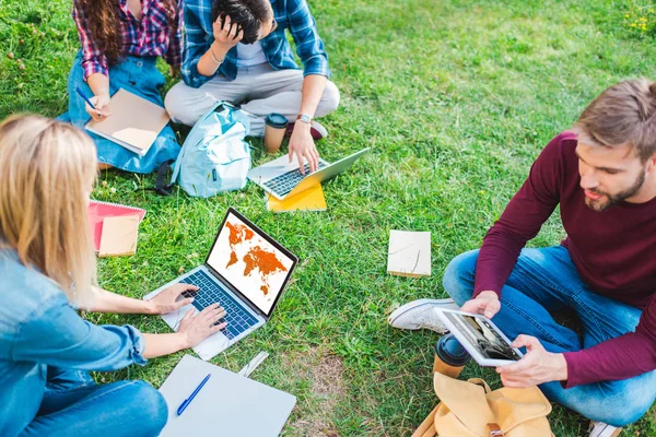 Partial view of multiethnic students with notebooks and digital devices sitting on green grass in park — Stock Photo