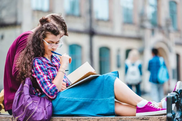 Partial view of young woman with book and notebook leaning on boyfriend while studying on wooden bench in park — Stock Photo