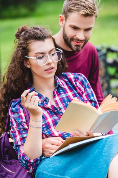 Ritratto di giovane donna con libro e quaderno appoggiato al fidanzato mentre studia su panchina in legno nel parco — Foto stock