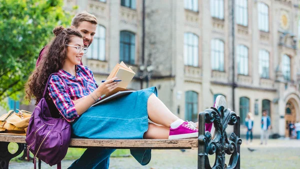 Усміхнена молода жінка з книгою і блокнотом спирається на хлопця на дерев'яній лавці в парку — стокове фото