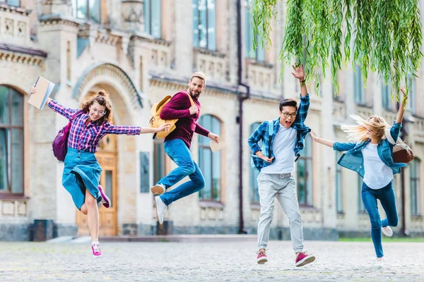 Happy multicultural students with backpacks jumping on street with university on background — Stock Photo