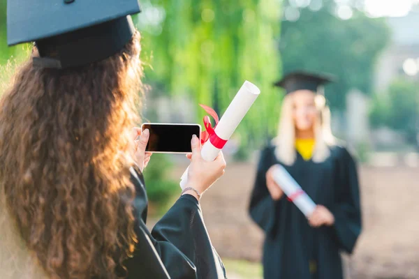 Vista parcial de la mujer tomando una foto de su compañero de clase en el teléfono inteligente durante la graduación cerca de la universidad - foto de stock