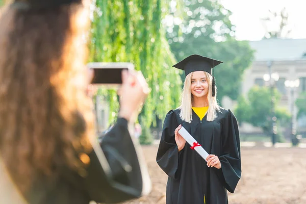 Vista parcial de la mujer tomando fotos de compañero de clase sonriente durante la graduación cerca de la universidad - foto de stock