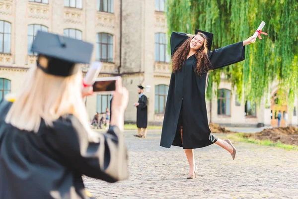 Vista parcial de la mujer tomando fotos de su compañero de clase con diploma durante la graduación cerca de la universidad - foto de stock