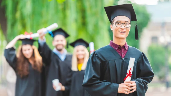 Selettivo fuoco di sorridente asiatico laureato con compagni di classe dietro in estate parco — Foto stock