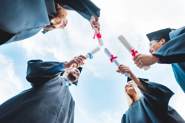 Bottom view of multiracial graduates with diplomas in hands and blue sky on background — Stock Photo