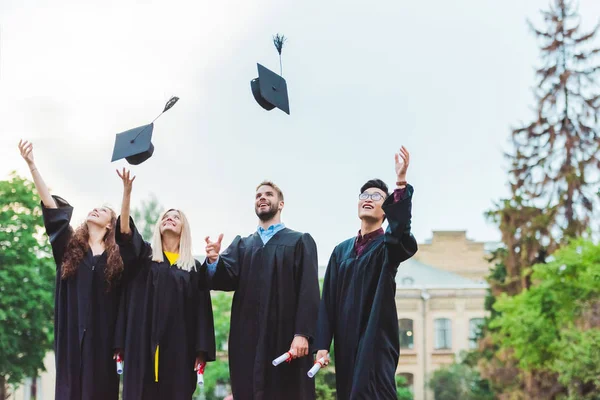 Porträt glücklicher multikultureller Absolventen mit Diplomen, die im Park Mützen hochwerfen — Stockfoto