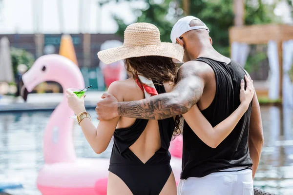 Вид ззаду хлопець і дівчина обіймаються і ходять біля басейну з коктейлем — стокове фото
