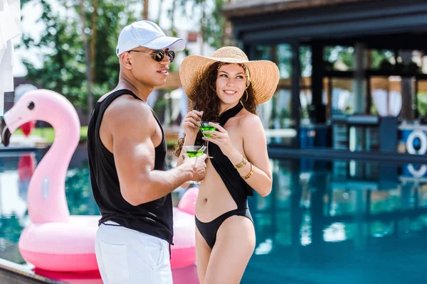 Smiling boyfriend and girlfriend standing with cocktails near swimming pool — Stock Photo