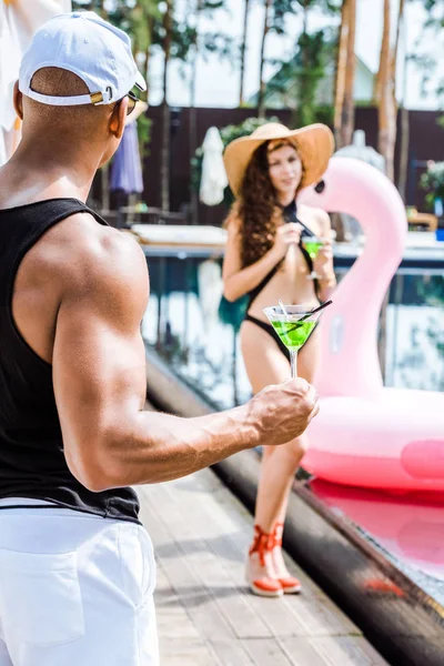 Freund hält Glas Cocktails in der Hand und schaut Freundin in Schwimmbadnähe an — Stockfoto
