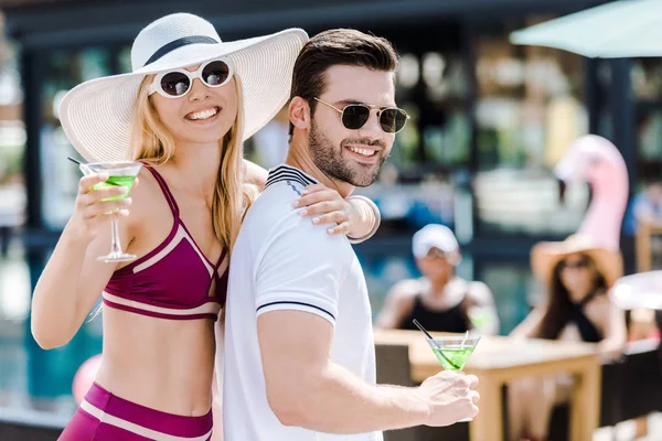Casal feliz em óculos de sol posando com copos de coquetéis perto da piscina — Fotografia de Stock