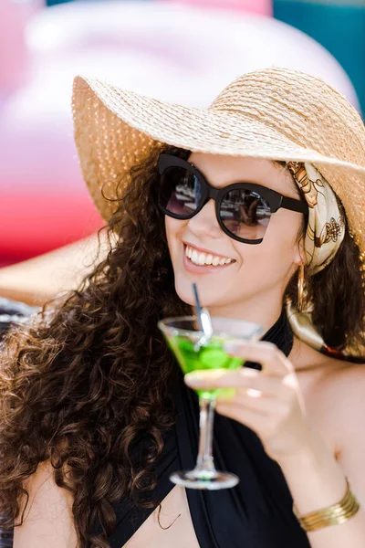 Atractiva chica sonriente en gafas de sol y sombrero de beber cóctel con paja - foto de stock