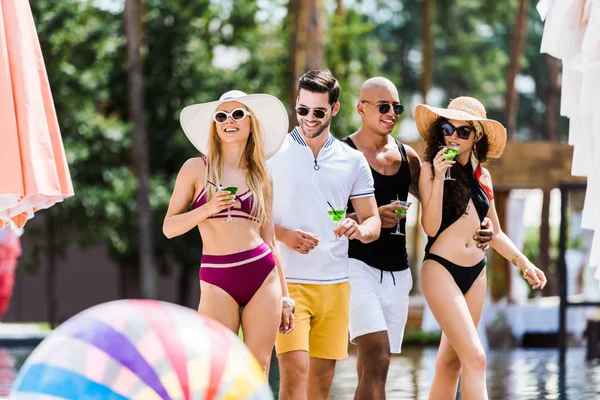 Four male and female friends walking near swimming pool with glasses of cocktails — Stock Photo