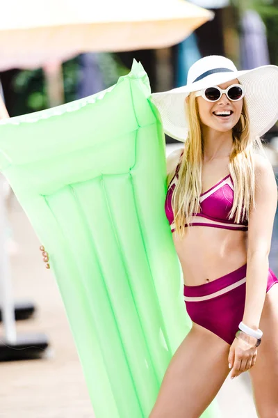 Sorridente ragazza attraente in costume da bagno e cappello in piedi con materasso gonfiabile vicino alla piscina — Foto stock