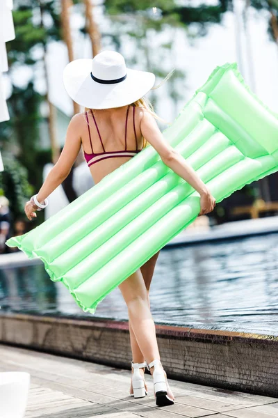 Rückansicht des Mädchens in Badeanzug und Hut, das mit aufblasbarer Matratze in der Nähe des Swimmingpools spaziert — Stockfoto