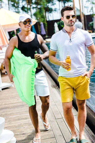 Bei amici maschi che camminano con materasso gonfiabile e birra vicino alla piscina — Foto stock