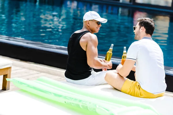 Amigos masculinos sonrientes sentados con cerveza cerca de la piscina - foto de stock