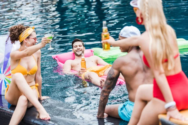 Sonriendo jóvenes amigos tintineo por botellas de cerveza y vaso de cóctel en la piscina - foto de stock