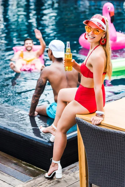 Улыбающаяся женщина, сидящая с пивом, пока ее друзья-мужчины развлекаются в бассейне — стоковое фото