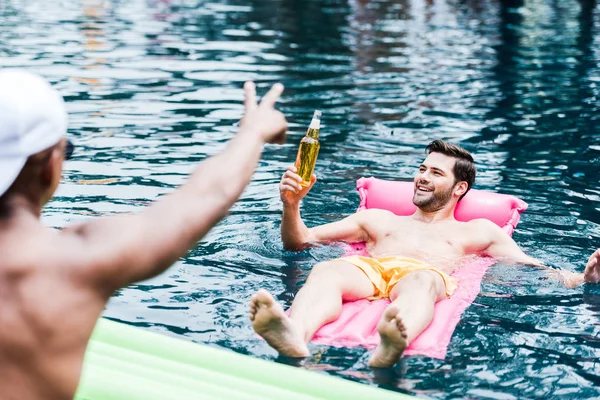 Homem sorridente com cerveja descansando no colchão inflável na piscina, enquanto seu amigo gesticulando por dedos na beira da piscina — Fotografia de Stock