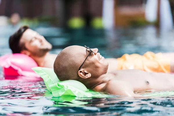 Amigos masculinos felices que descansan y que toman el sol en colchones inflables en piscina - foto de stock