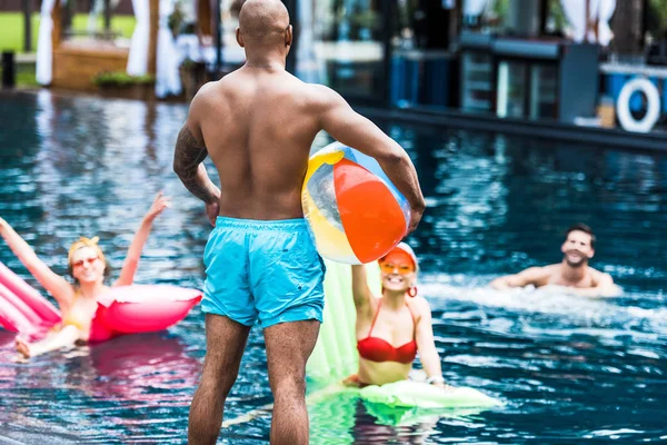 Rear view of man standing with swimming ball while his friends in swimming pool with inflatable mattresses — Stock Photo