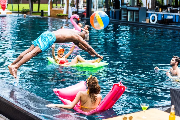 Junger Mann springt ins Schwimmbad, während seine Freundinnen sich auf aufblasbaren Matratzen ausruhen — Stockfoto