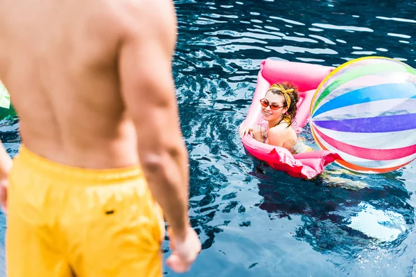 Imagen recortada del hombre de pie en la piscina mientras que su novia nadando en colchón inflable con bola de natación en la piscina - foto de stock