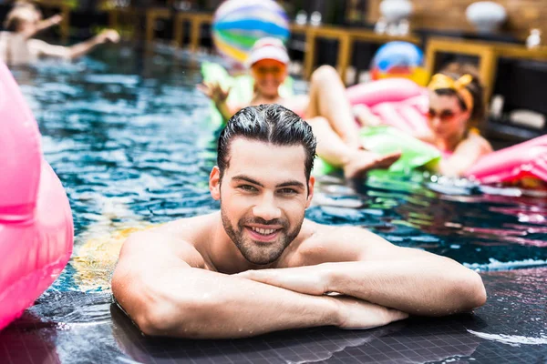 Jovem feliz olhando para a câmera perto da piscina, enquanto seus amigos do sexo feminino descansando em colchões infláveis na piscina — Fotografia de Stock