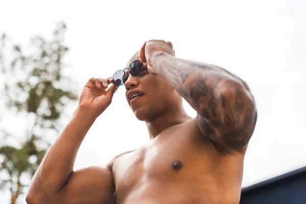 Низкий угол зрения на мускулистого татуированного человека, снимающего солнцезащитные очки — стоковое фото