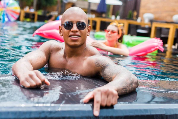 Joven hombre tatuado en gafas de sol mirando a la cámara mientras su novia descansa sobre un colchón inflable en la piscina - foto de stock