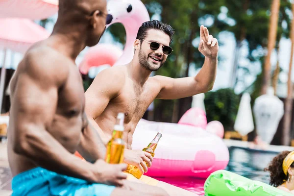 Sorrindo homem em óculos de sol gesticulando à mão, enquanto seu amigo sentado perto com cerveja na beira da piscina — Fotografia de Stock