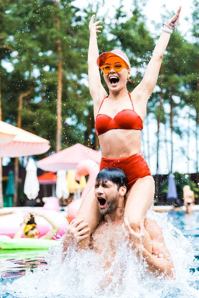 Heureuse jeune femme avec les bras levés assis sur les épaules de copains dans la piscine — Photo de stock