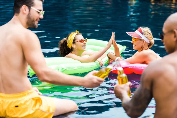 Sorrir amigos do sexo feminino tendo alta cinco para o outro na piscina, enquanto amigos do sexo masculino clinking por garrafas de cerveja — Fotografia de Stock