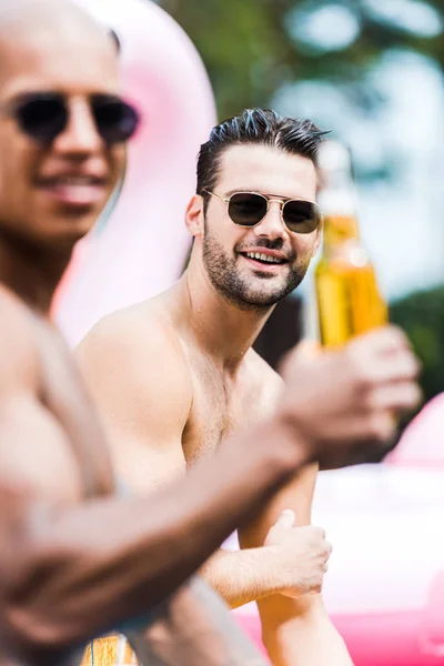 Усміхнений чоловік в сонцезахисних окулярах дивиться на камеру, а його друг сидить поруч з пивом — стокове фото