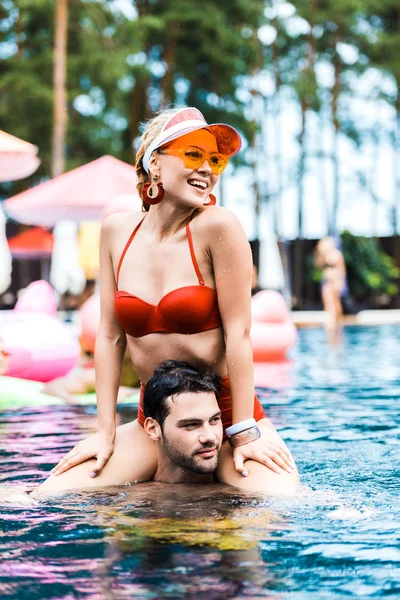 Mujer sonriente en traje de baño rojo mirando hacia otro lado mientras se sienta sobre los hombros de los novios en la piscina - foto de stock