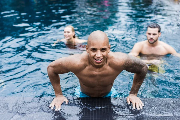 Joven tatuado mirando a la cámara mientras sus amigos nadan en la piscina detrás - foto de stock