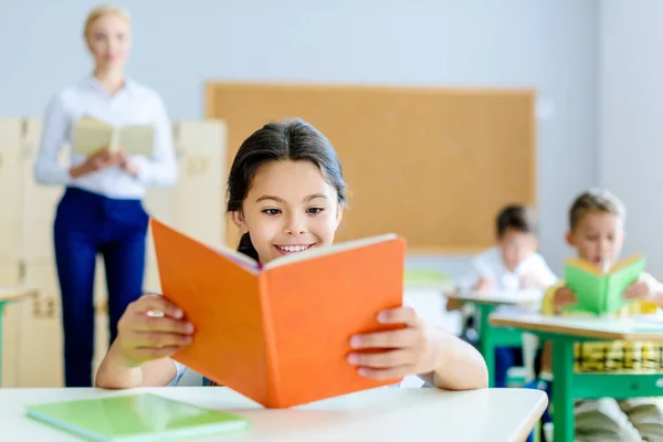 Beau sourire écolière lecture livre à salle de classe pendant la leçon — Photo de stock