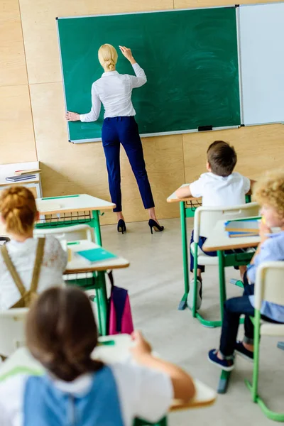 Vue arrière des enfants regardant le professeur alors qu'elle écrit sur un tableau — Photo de stock