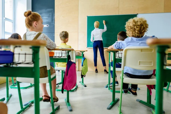 Vista trasera de los escolares mirando a la maestra mientras escribe en pizarra - foto de stock