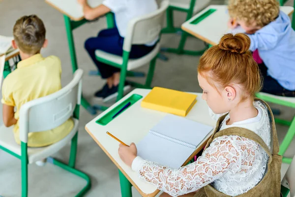 Belle écolière rousse écrivant dans un cahier pendant la leçon — Photo de stock