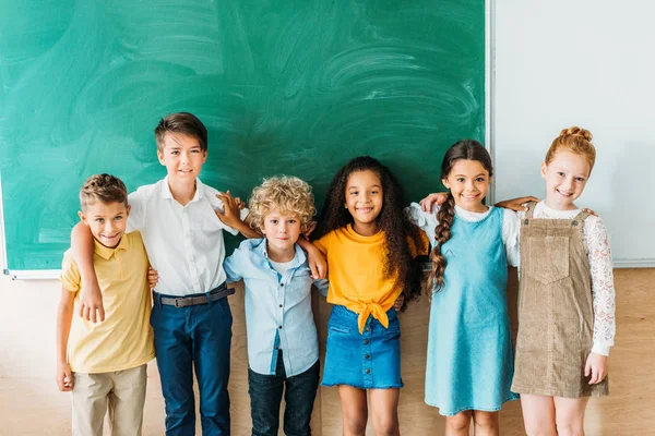 Group of multiethnic schoolchildren embracing in front of blank chalkboard — Stock Photo