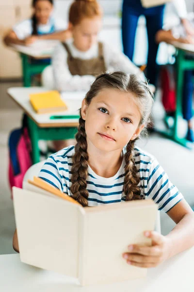 Schöne kleine Schülerin mit Buch, die während des Unterrichts in der Schule in die Kamera schaut — Stockfoto