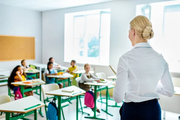 Vista trasera del profesor mirando a los niños sentados en el aula - foto de stock