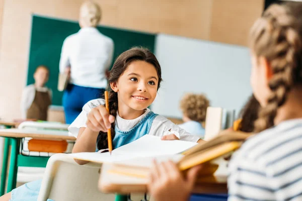 Счастливая маленькая школьница разговаривает с одноклассницей во время урока — стоковое фото
