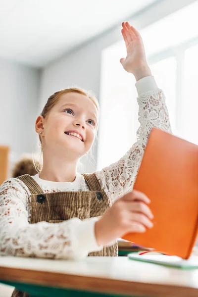Sorridente scolaretta con libro alzando la mano durante la lezione — Foto stock