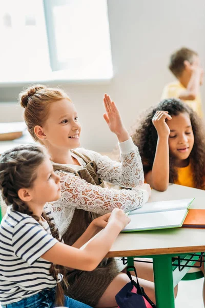 Meninas felizes sentadas na sala de aula durante a aula e levantando as mãos — Fotografia de Stock