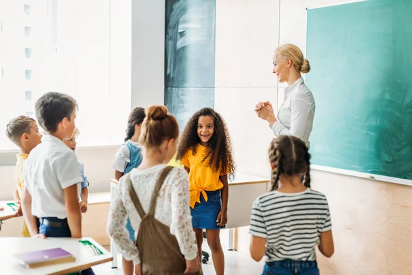 Compañeros de clase multiétnicos felices de pie alrededor del profesor en el aula - foto de stock