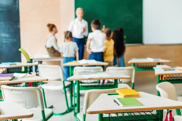 Schoolchildren standing around teacher at classroom with desks on foreground — Stock Photo